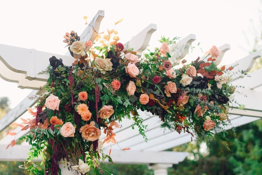 Large floral installation featuring peach roses, coral dahlias, burgundy foliage and cascading greenery arranged across a white pergola at an outdoor wedding ceremony.